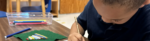 A young boy holding a pencil and doing work with some Montessori material in the background